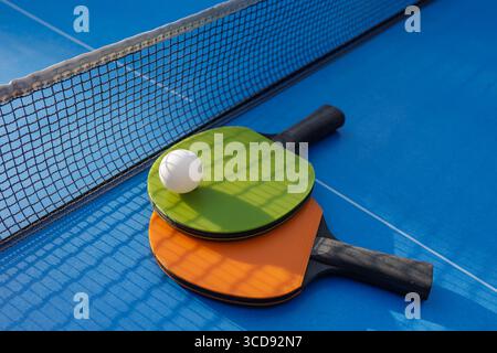 Sportliches Tischtennis-Stillleben mit Ball auf bunten Paddles am Netz, blauen Schatten auf dem Platz und Kopierraum für Training, Schule oder Wettkampf Stockfoto