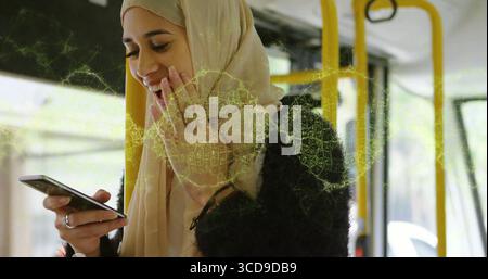Lächelnde Frau mit Hijab und schwarzer Jacke, Telefon im Bus an Fenstern mit gelben Stangen Stockfoto
