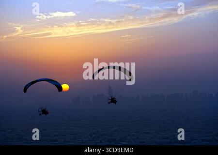 Zwei Paramotoren fliegen bei Sonnenuntergang entlang der Küste, während sich die ferne Stadt Skyline im Abendlicht abzeichnet. Stockfoto