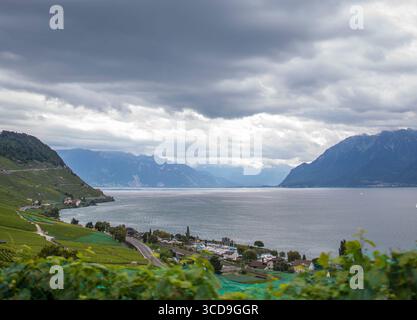 Malerischer Blick auf den Genfer See mit dramatischem bewölktem Himmel, umliegenden Weinbergen, Bergen und friedlichem Dorf am Ufer in der Schweiz. Stockfoto