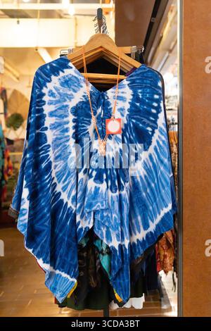 Ein leuchtend blauer Poncho mit Bindegürtel befindet sich in einem stilvollen Modegeschäft auf einem Holzbügel und zeigt komplizierte Designs. Stockfoto