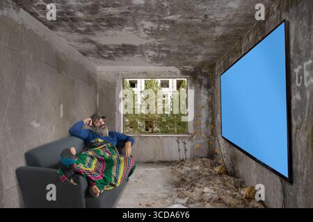 Bärtiger Mann, der auf der Couch in einem verlassenen Zimmer liegt und an der Wand mit blauem Bildschirm fernsieht Stockfoto