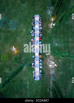 Blick aus der Vogelperspektive auf einen langen Esstisch inmitten von üppigem grünem Gras, Bäumen und verstreuten Leuchten, Assisi, Umbrien, Italien. Stockfoto
