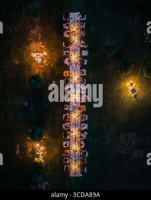 Blick aus der Vogelperspektive auf einen langen Tisch für ein warmes, einladendes Abendessen unter dem sanften Licht im dunklen, grünen Rasen, Assisi, Umbrien, Italien. Stockfoto