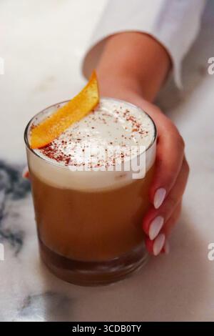 Hand hält einen Craft-Cocktail in einem kurzen Glas, gekrönt mit einer cremigen Schaumschicht, staubtem Gewürz und garniert mit einer frischen Orangenschale Stockfoto