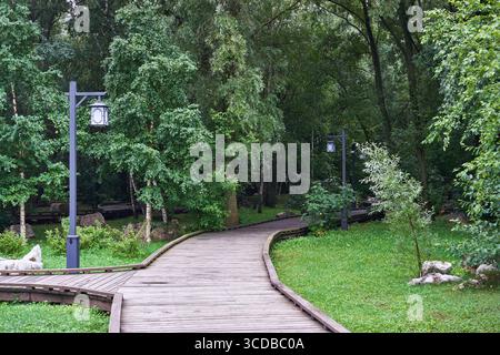 Moskau, Russland - 17. Juli 2025: Kurviger Holzsteg mit Laternen durch üppige grüne Bäume in einem ruhigen Stadtpark Stockfoto