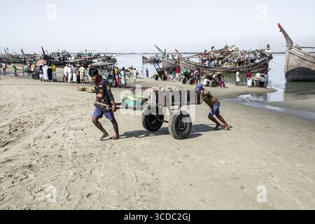 Cox's Bazar, Bangladesch - 29. November 2022: Blick auf Männer, die einen schweren Wagen durch den Sandstrand ziehen wollen, mit Fischerbooten, die sich unter einem trüben Himmel an der Küste ansammeln. Stockfoto
