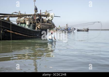 Cox's Bazar, Bangladesch - 29. November 2022: Blick auf einen einsamen Fischer, der sein Netz in den glitzernden Gewässern wirft, traditionelle Boote, die unter klarem Himmel am Horizont liegen. Stockfoto