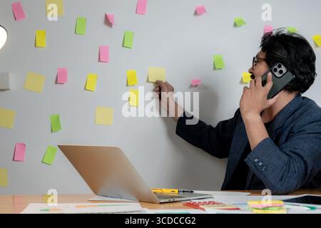 Ein junger Geschäftsmann schreibt Ideen auf Haftnotizen und klebt sie an einer Wand in seinem Büro. Stockfoto