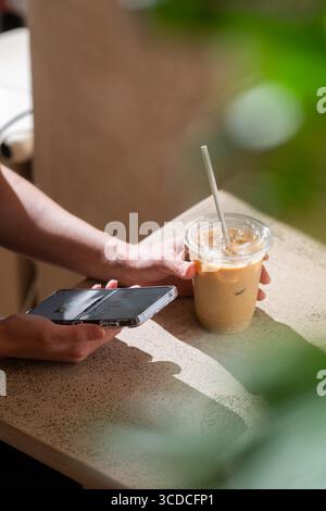 Nahaufnahme einer Frau, die ein Smartphone in der einen Hand hält und in der anderen ein Glas Eiskaffee in einem modernen Café. Stockfoto