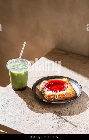 Ein erfrischender Matcha-Latte, serviert mit Erdbeergebäck auf einem Teller, der auf einem minimalistischen Coffee-Shop-Tisch serviert wird. Stockfoto