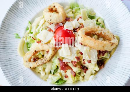 Farbenfroher Salat im Caesar-Stil wird mit knusprigen Calamari-Ringen, Kirschtomaten, Speckstückchen und geriebenem Parmesan belegt Stockfoto