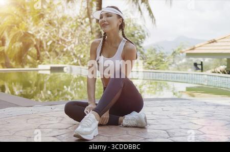 Ich war begeistert, dass die schlanke Dame in Sportkleidung lächelte und wegschaute, während sie am sonnigen Tag im tropischen Resort in der Nähe des Swimmingpools saß Stockfoto