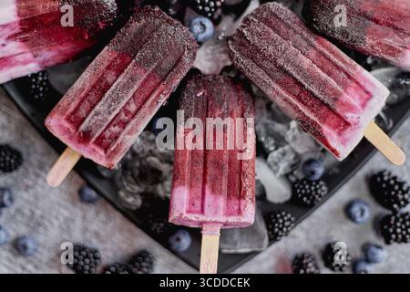 Hausgemachte frisch gefrorene Heidelbeere und Brombeerpopsicles auf schwarzem Teller mit Eis sitzen auf Stein Tisch mit frischen Beeren auf der Seite. Draufsicht, flach liegend Stockfoto