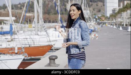 Attraktive, lässige junge Frau aus Denim, die an einer Uferpromenade steht und über etwas links vom Rahmen lacht Stockfoto