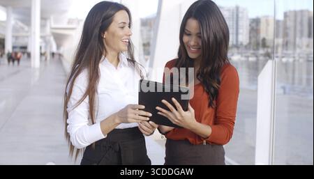 Zwei fröhlich lächelnde, elegant gekleidete Geschäftsfrauen sprechen miteinander über ihr Projekt auf einem Tablet-Gerät Stockfoto