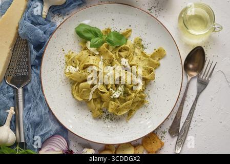 Leckere Tagliatelle-Pasta mit Basilikum und grünem Pesto, serviert auf Keramikplatte mit Zutaten an den Seiten. Draufsicht, flach Stockfoto