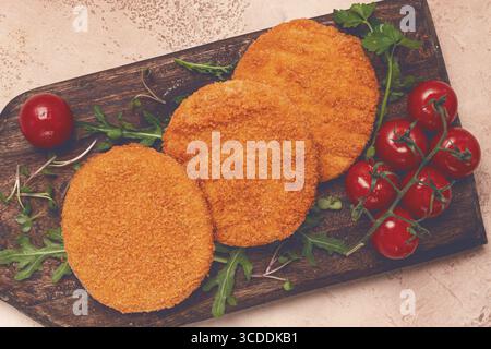 Knusprig gebratene Fischkoteletts, Fischburger, auf einem Holzschneidebrett, mit Rucola und Kirschtomaten, hausgemacht, keine Leute Stockfoto