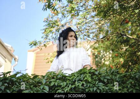 Eine Frau mit dunklen, lockigen Haaren steht inmitten üppig grüner Blätter und blickt nachdenklich in die Ferne. Der hellblaue Himmel über ihnen verstärkt ihr heiteres P Stockfoto