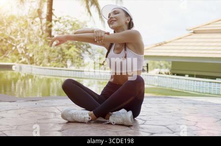 Glückliche schlanke Dame in Sportbekleidung, die Arme mit geschlossenen Augen dehnt, während sie beim Training am sonnigen Tag im Resort mit überkreuzten Beinen am Pool sitzt Stockfoto