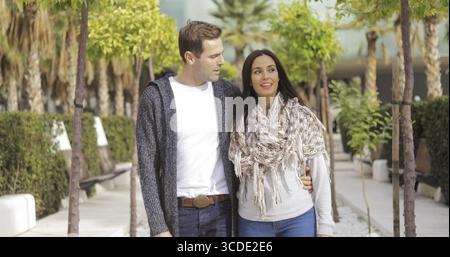 Trendige zärtlich junges Paar gehen Arm in Arm entlang einer Promenade zwischen Setzlingen in einem städtischen Park, plaudern und lachen Stockfoto