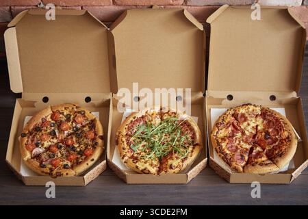 Drei verschiedene Arten von Pizzen in Lieferboxen. In einer Reihe auf braunem hölzernem Hintergrund platziert. Ansicht von oben mit einem Leerzeichen für Ihren Text Stockfoto