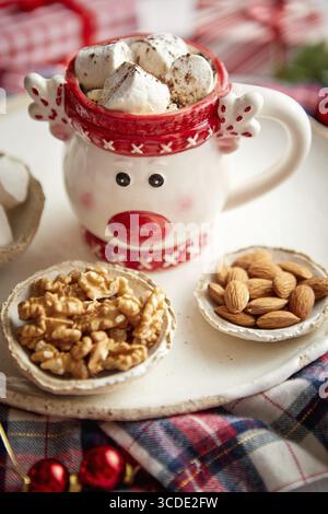 Köstliche hausgemachte heisse weihnachtsschokolade oder Kakao mit Marshmellows in einem roten Weihnachtsbecher. Mit Mandeln, Walnüssen in kleinen Schüsseln an der Seite Stockfoto