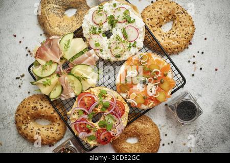 Köstliche Bagel Sandwiches mit Käse, Schinken, Hummus, Lachs und Gemüse. Serviert auf schwarzem Grill auf weißem rustikalem Tisch. Gesundes hausgemachtes Essen Stockfoto