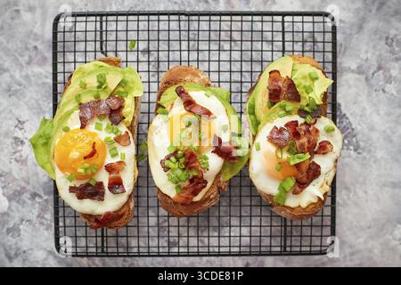 Leckere hausgemachte Toasts mit Spiegelei, Speck, Avocado, Salat und Schnittlauch. Serviert auf schwarzem Grill Stockfoto