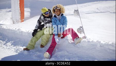 Glückliches junges Paar in farbenfrohen Skikleidung, das sich auf einem Schelfeige aus frischem, weißem Winterschnee entspannt und lachend in der Sonne sitzt Stockfoto