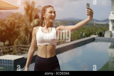 Glückliche schlanke Frau in Sportkleidung lächelt und macht Selfie gegen den Swimmingpool während des Fitnesstrainings an sonnigen Tagen im Resort Stockfoto
