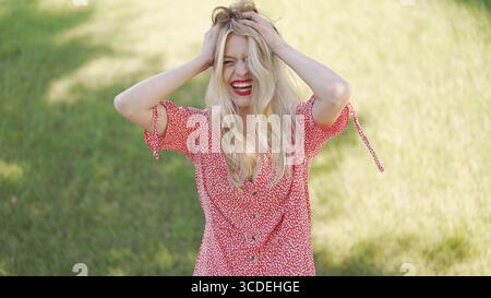 Eine fröhliche junge Frau in einem roten Blumenkleid steht auf einem sonnendurchfluteten Feld und strahlt ein Gefühl von Freude und Freiheit aus Stockfoto