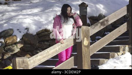 Entzückendes asiatisches Mädchen, das auf Holztreppen im schneebedeckten Bergresort steht. Sie trägt Snowboarderkleidung Stockfoto