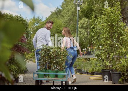 Männliche und weibliche kooperierende professionelle Gärtner, die gemeinsam einen Wagen mit Topfblumen im Garten tragen Stockfoto