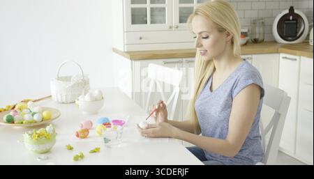 Junge schöne Blondine in lässigem Outfit, die am Tisch in der Küche sitzen und Eier für die Osterferien malen Stockfoto