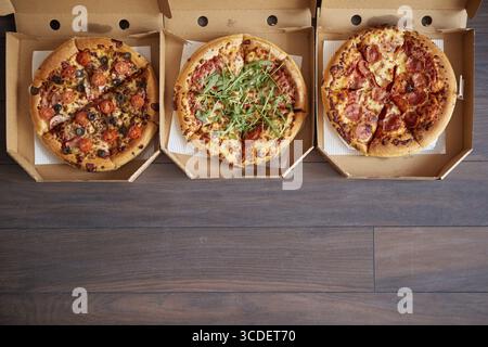 Drei verschiedene Arten von Pizzen in Lieferboxen. In einer Reihe auf braunem hölzernem Hintergrund platziert. Ansicht von oben mit einem Leerzeichen für Ihren Text Stockfoto