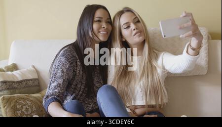 Zwei hübsche Mädchen sitzen zusammen auf einem Sofa im Wohnzimmer und posieren für ein Selfie auf einem Handy lächelnd und lachend Stockfoto