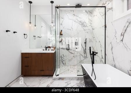 Ein Badezimmer mit Wänden und Dusche aus Calacatta-Marmor, Hängelampen über dem weißen Waschbecken und Holzschränke und eine schwarz-weiße Badewanne. Stockfoto