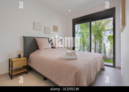 Ruhiges Schlafzimmer mit zartem rosafarbenem Bettzeug, Tageslicht und einer großen Schiebetür, die zu üppigem Grün führt. Stockfoto