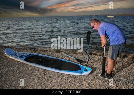 Ein Mann, der sein Stehpaddelbrett an einem Kieselstrand bei Sonnenuntergang aufbläst und sich auf Wassersport vorbereitet. Stockfoto
