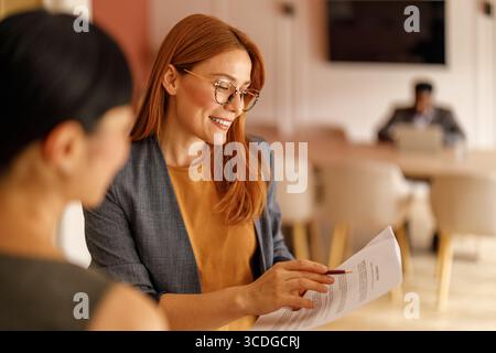 Lächelnde Anwältin in Brille, die der Geschäftsfrau beim Gespräch im Amt Vertragspapiere zeigt Stockfoto
