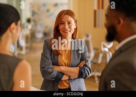 Erfolgreiche Unternehmerin mit überkreuzten Armen lächelnd in die Kamera, während sie sich mit Kollegen im Meeting besprach Stockfoto