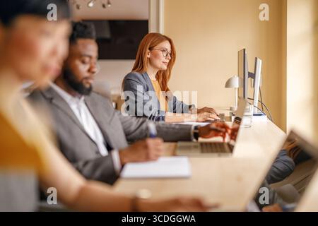 Fokussierte Geschäftsfrau in Brille, die am Computer arbeitet, während sie mit Kollegen am Schreibtisch in einem modernen Büro sitzt Stockfoto