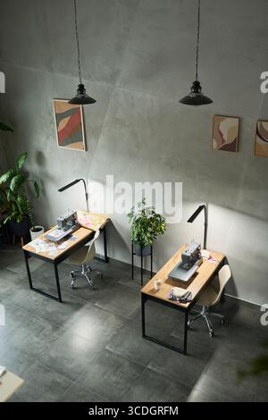 Leerer moderner Arbeitsplatz mit zwei Nähmaschinen auf Holzschreibtischen mit Stoffstücken, ergonomischen Stühlen, Topfpflanzen, abstrakten Wandbildern, Hängeleuchten, gefliester Boden Stockfoto