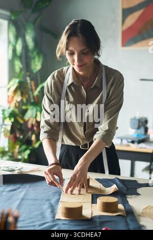 Junge Erwachsene kaukasische Frau, die mit Nähmustern auf Stoff arbeitet, Material in der Werkstatt misst und schneidet, Band um den Hals, konzentriert sich auf den Prozess der Bekleidungsherstellung Stockfoto