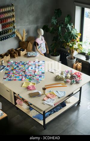 Arbeitstisch aus Textil mit bunten Stoffquadraten, Nähwerkzeugen, Papiermustern und Stoffresten, Schaufensterpuppe mit Patchworkschürze neben Fenster, Fadenspulen an der Wand Stockfoto