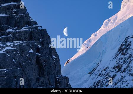 Ein Halbmond erhebt sich im Abendlicht über schneebedeckten Gipfeln und Gletschern im Lemaire-Kanal, der Antarktischen Halbinsel, der Antarktis. Stockfoto