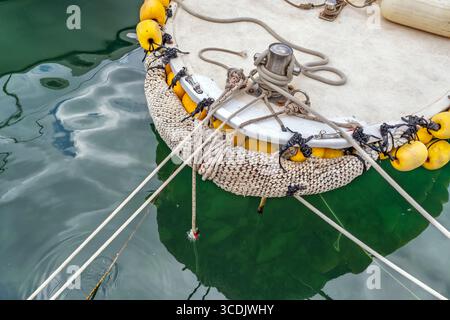 Nahaufnahme des Bugs eines Bootes mit Anlegeseilen, einer Stange und einem traditionellen gewebten Kotflügel mit gelben Bojen, der in einem Hafen angedockt ist. Nautische Details Stockfoto