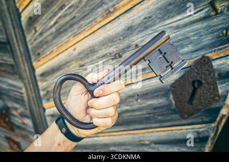 Ein großer, altmodischer Schlüssel wird in der Hand vor einer verwitterten Holztür mit einer rostigen Schlüssellochplatte gehalten. Stockfoto