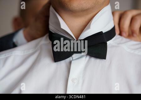 Ein Nahaufnahme-Porträt zeigt einen Bräutigam, der sich bereit macht, jemand, der seine schwarze Fliege auf einem knusprigen weißen Hemd anpasst, was Eleganz, Vorbereitung und symbolisiert Stockfoto